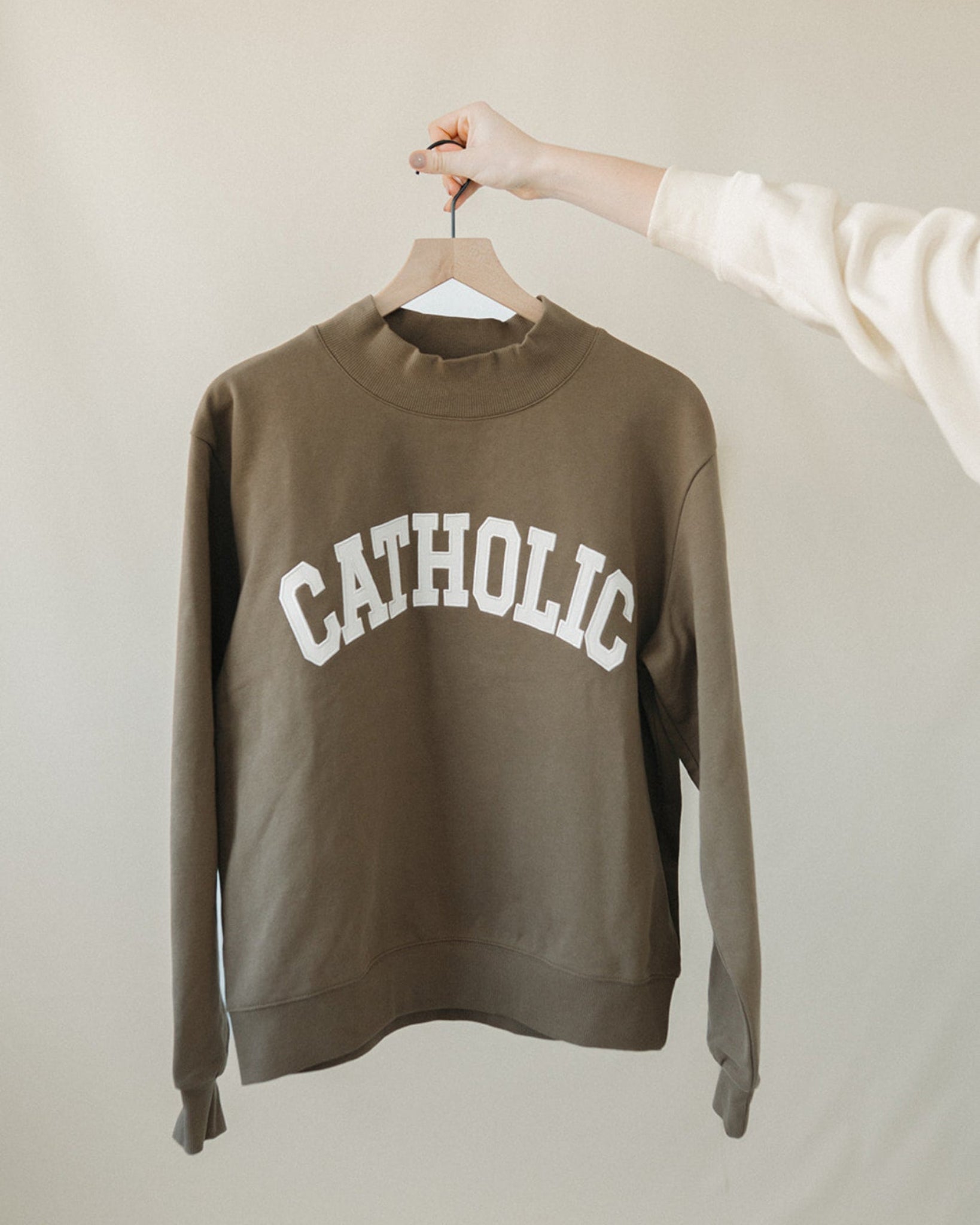 Green sweatshirt with 'CATHOLIC' text on a hanger against a beige background