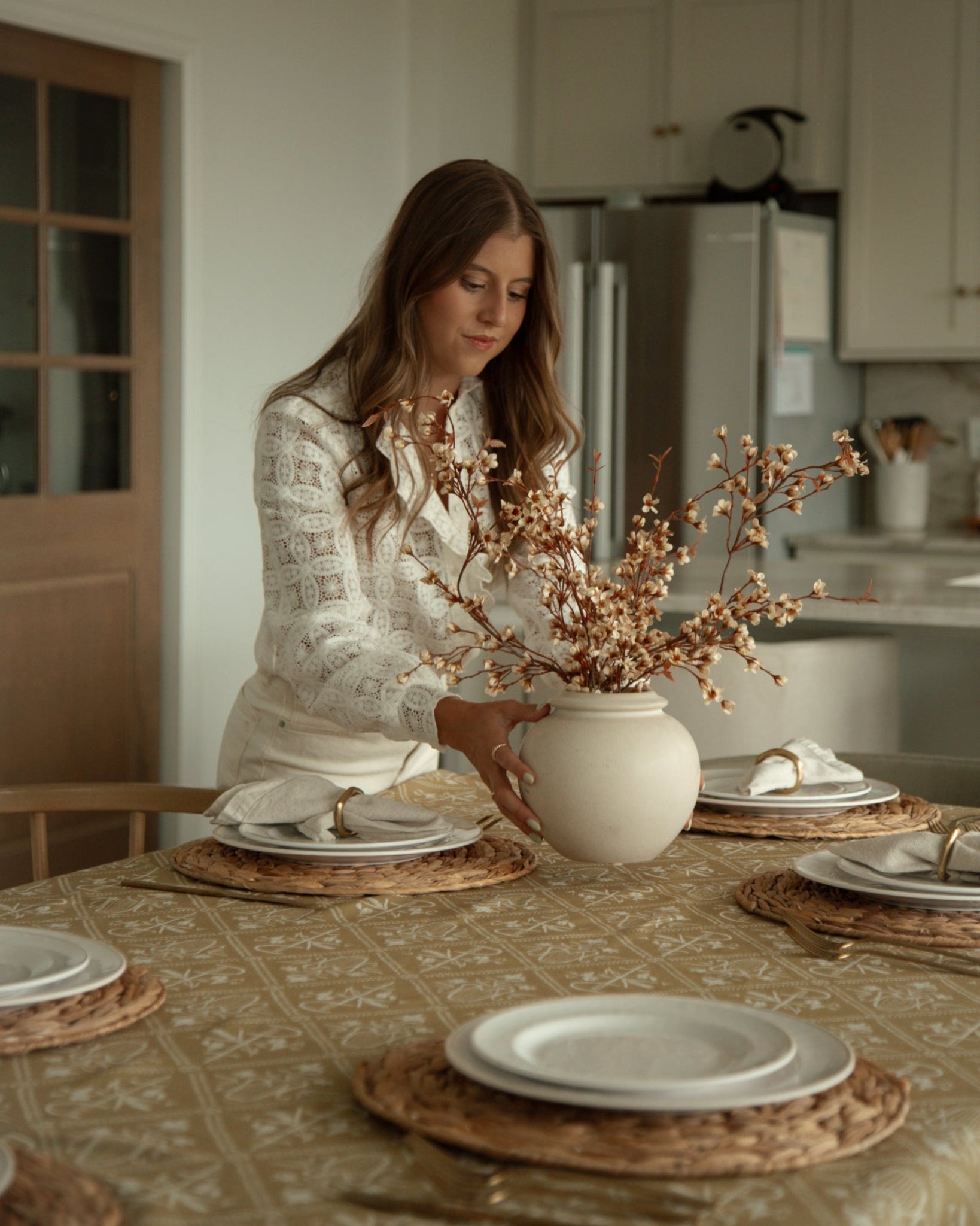 A woman in a white patterned blouse stands at a dining table in a bright kitchen, holding a white ceramic vase filled with dried beige branches. The table is set with white plates, white napkins, woven placemats and a Catholic inspired yellow tablecloth.