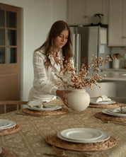 A woman in a white patterned blouse stands at a dining table in a bright kitchen, holding a white ceramic vase filled with dried beige branches. The table is set with white plates, white napkins, woven placemats and a Catholic inspired yellow tablecloth.