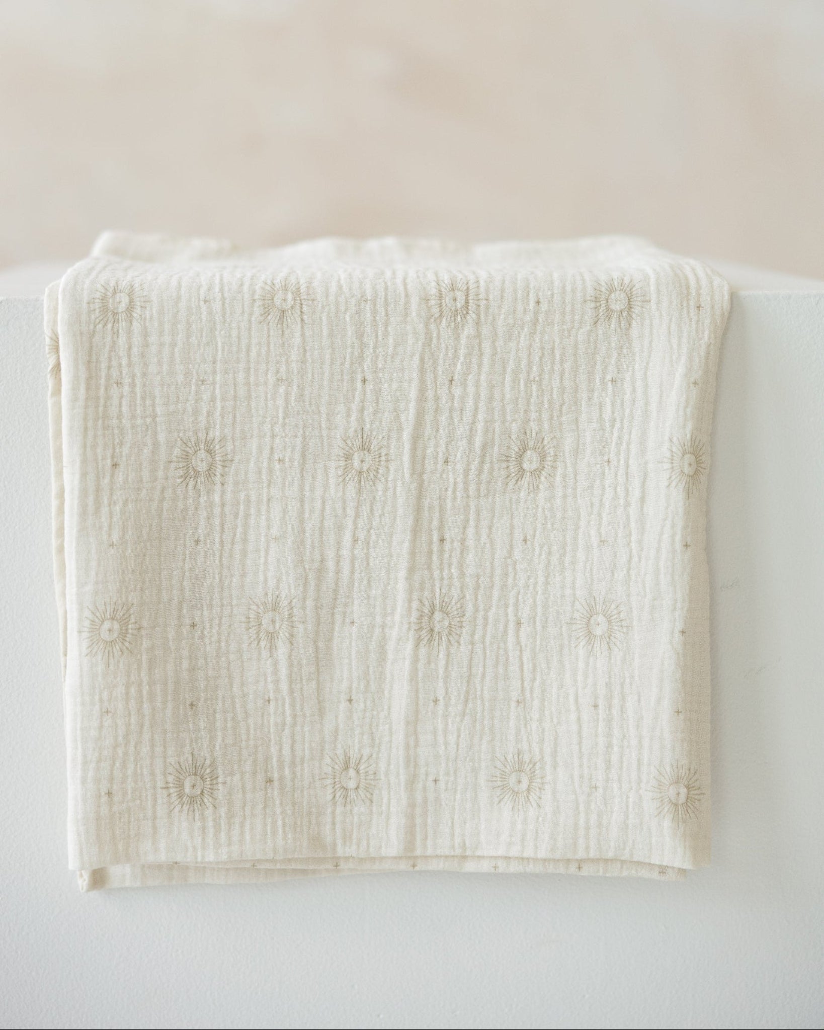 An off-white cotton muslin blanket with a textured crinkle weave and small embroidered floral motifs, draped over a white surface against a soft beige background.