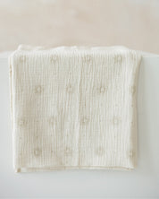 An off-white cotton muslin blanket with a textured crinkle weave and small embroidered floral motifs, draped over a white surface against a soft beige background.