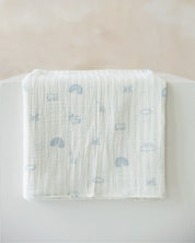 A folded white muslin baby blanket with a light blue pattern of rainbows, clouds, and animals, resting on a white surface against a soft beige background.