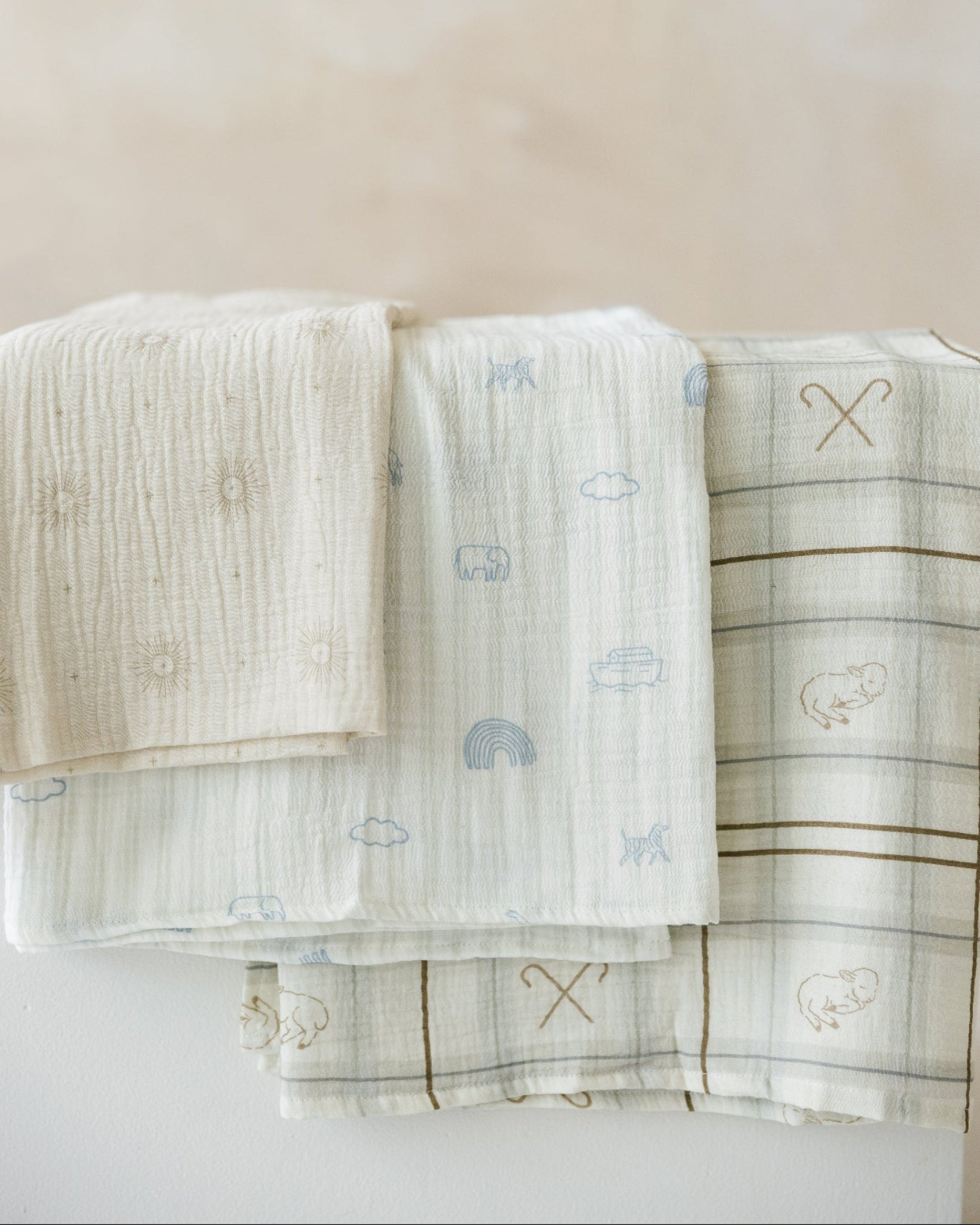 Three folded muslin baby blankets in soft neutral tones with printed motifs, arranged on a light surface against a warm beige background.