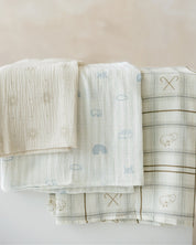 Three folded muslin baby blankets in soft neutral tones with printed motifs, arranged on a light surface against a warm beige background.