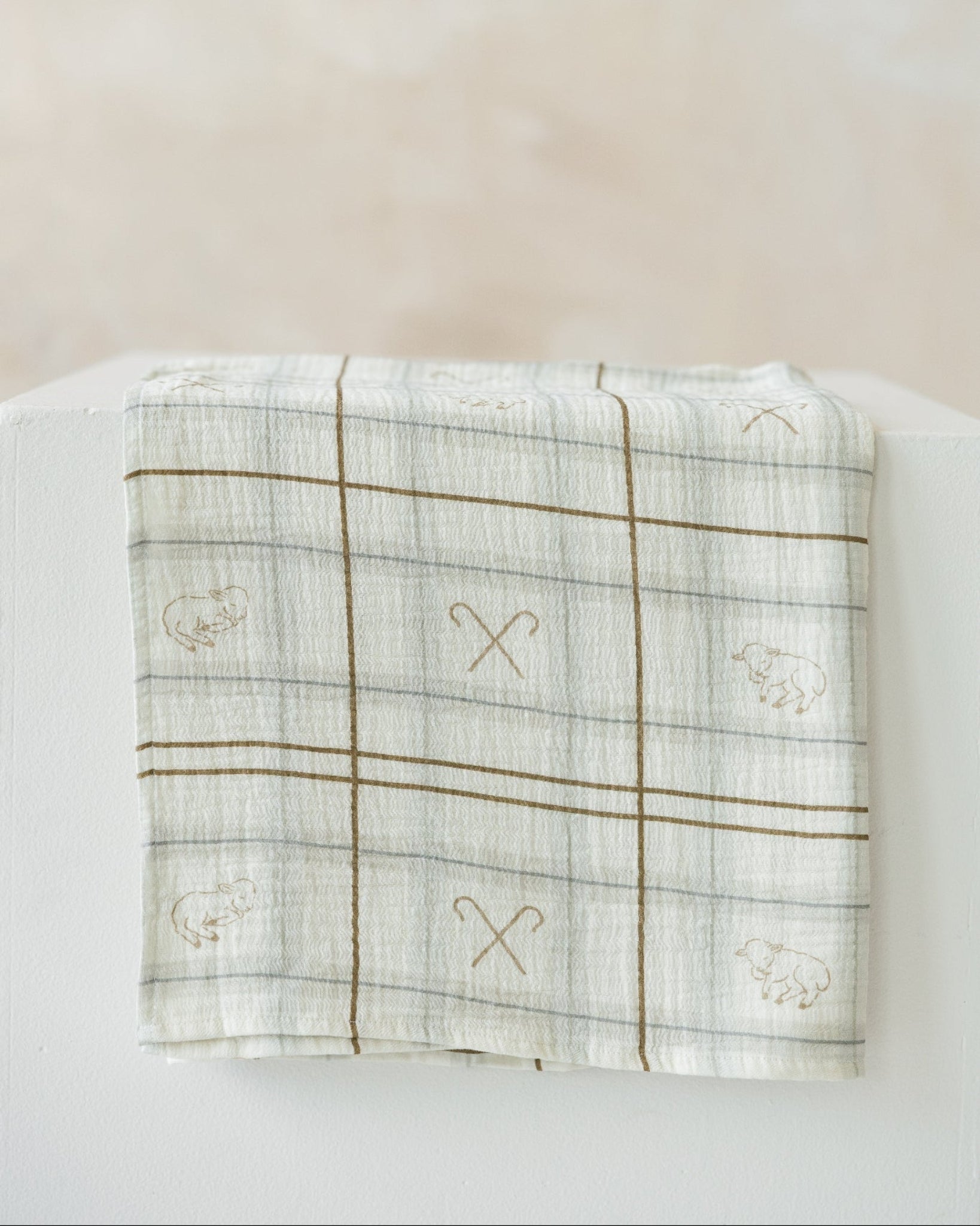 A folded white muslin baby blanket with a brown plaid grid pattern and embroidered sheep motifs, displayed on a white block against a soft beige background.