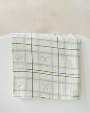 A folded white muslin baby blanket with a brown plaid grid pattern and embroidered sheep motifs, displayed on a white block against a soft beige background.
