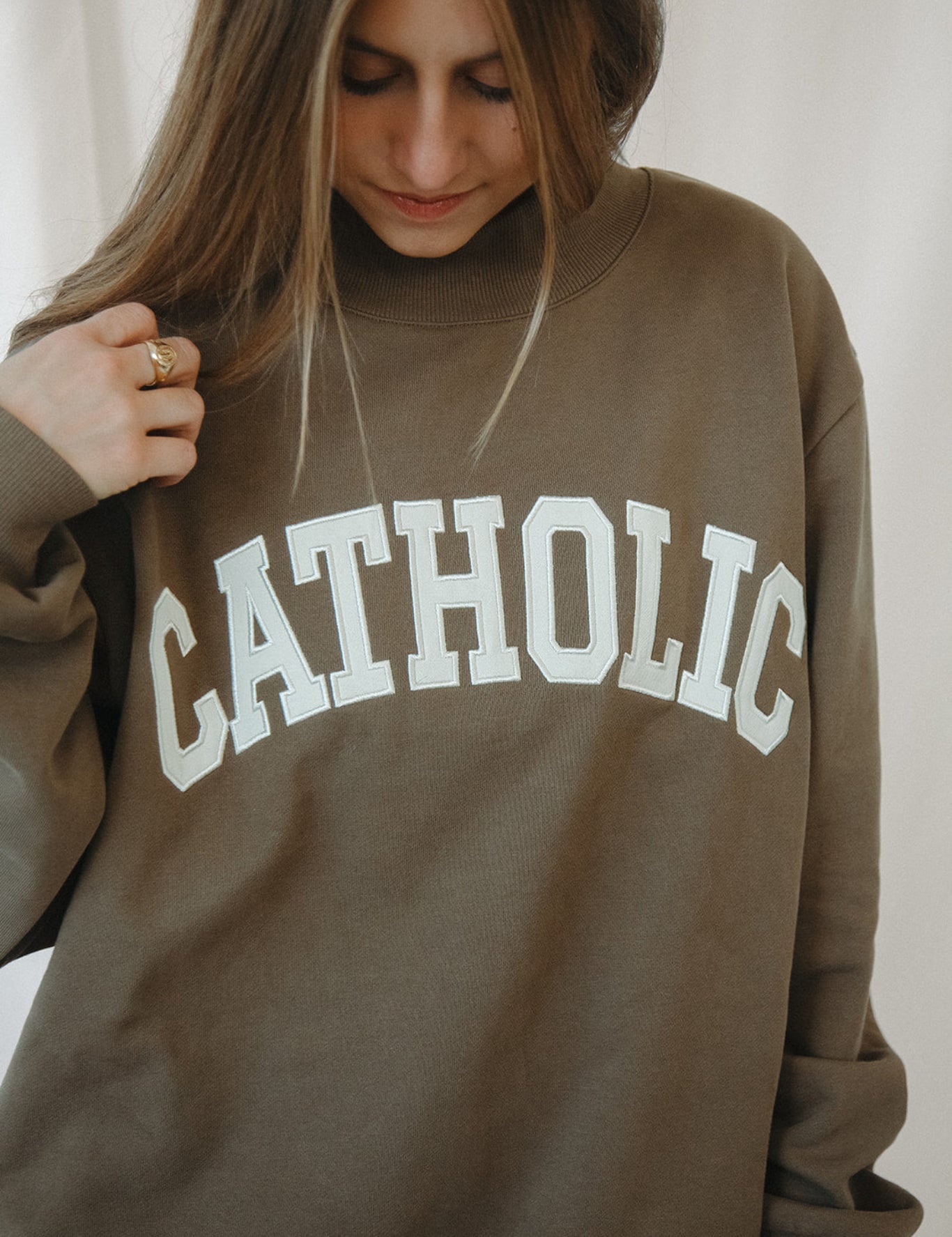 Person wearing a brown mockneck sweatshirt with 'CATHOLIC' printed on it.
