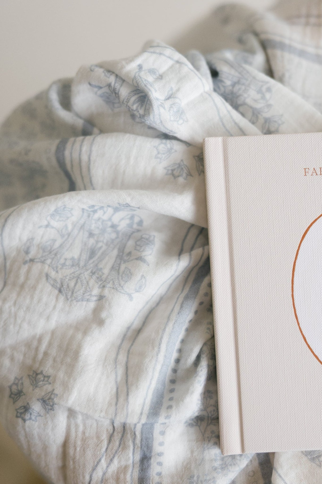 A close-up of a soft, textured baby blanket with a muted blue floral, Marian symbols and stripe pattern, draped over a cream-colored baby book. 