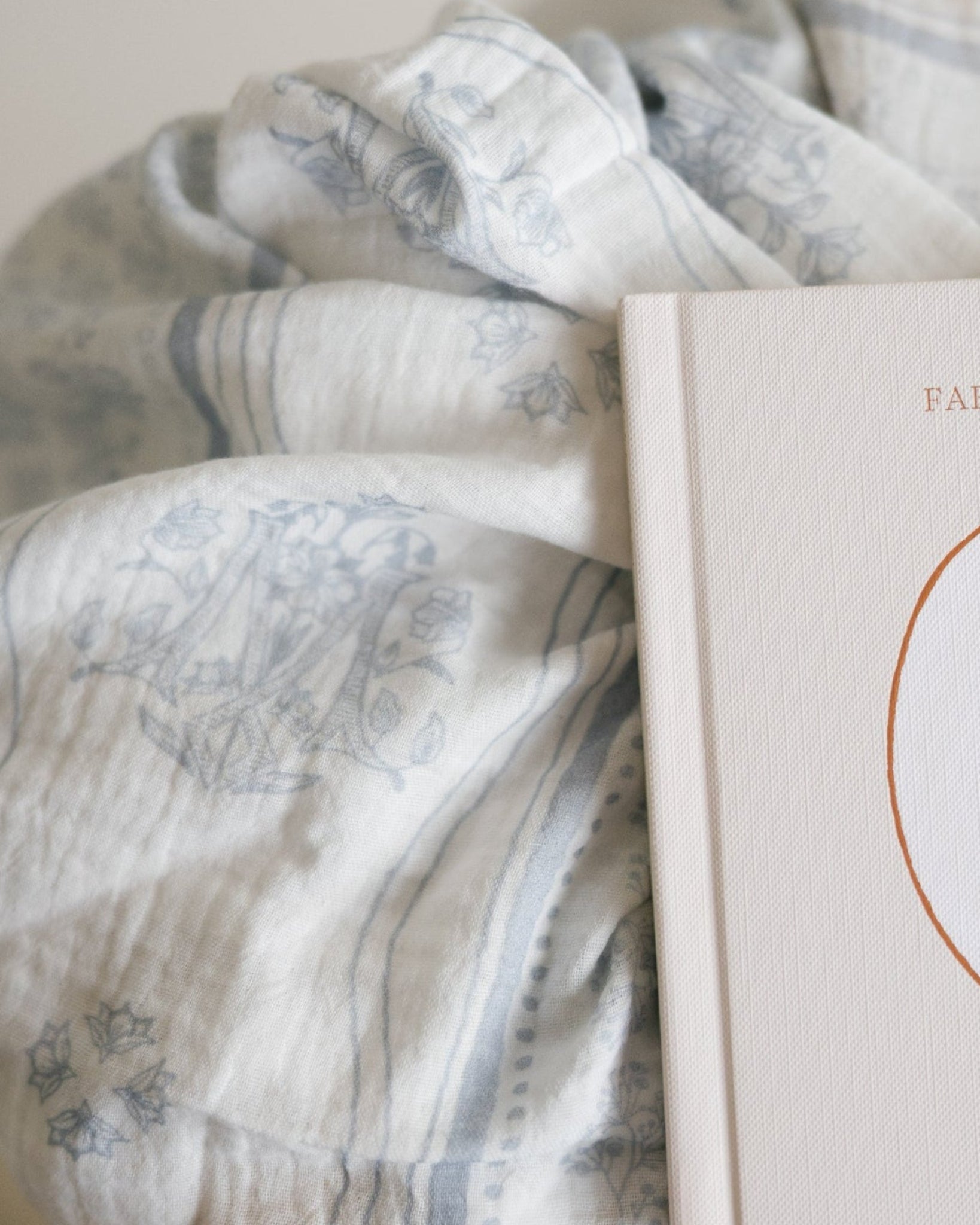 A close-up of a soft, textured baby blanket with a muted blue floral, Marian symbols and stripe pattern, draped over a cream-colored baby book. 