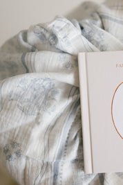 A close-up of a soft, textured baby blanket with a muted blue floral, Marian symbols and stripe pattern, draped over a cream-colored baby book. 