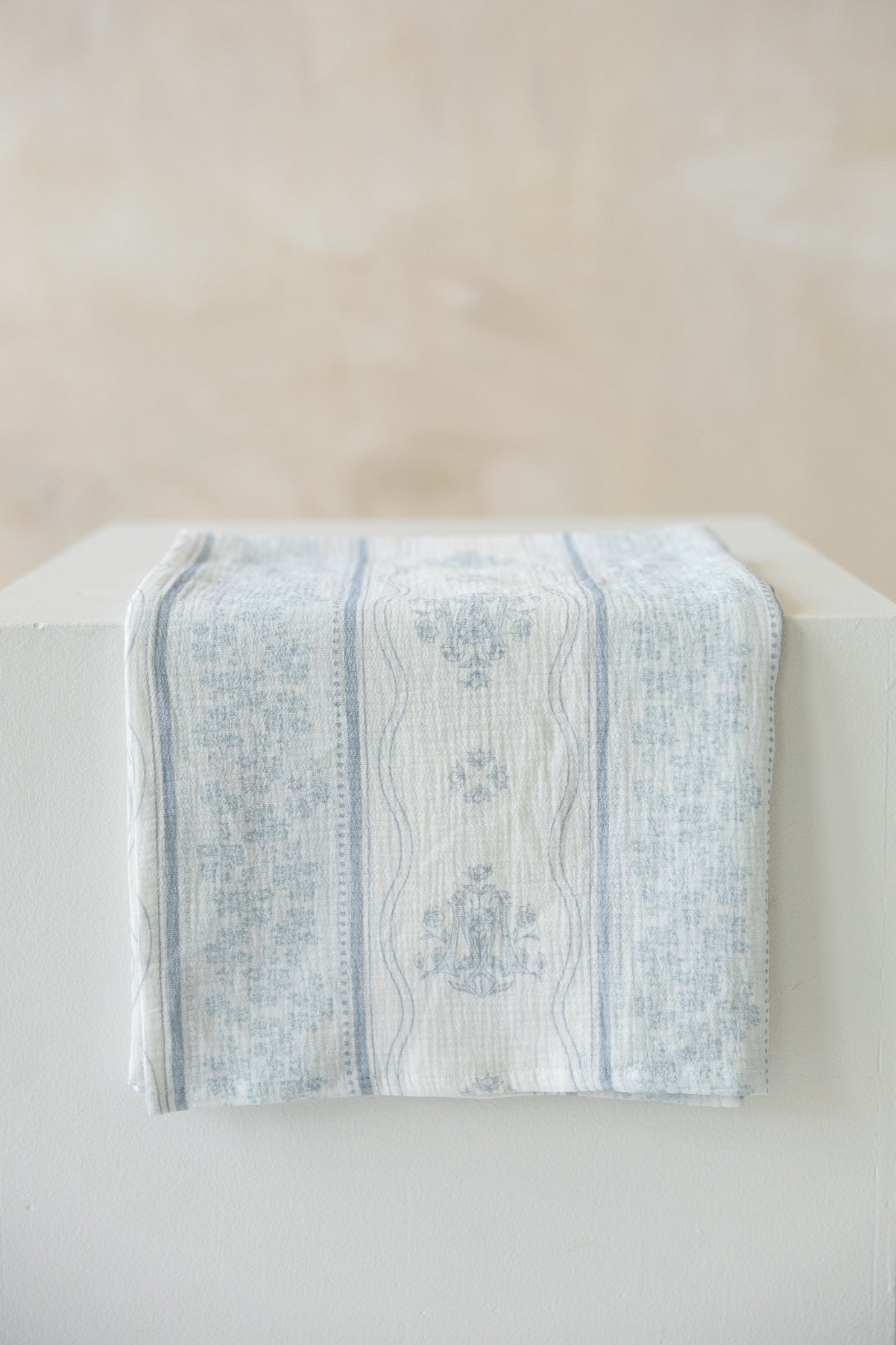 A light blue and white Marian and floral patterned cotton muslin cloth draped over a white block against a neutral beige background.