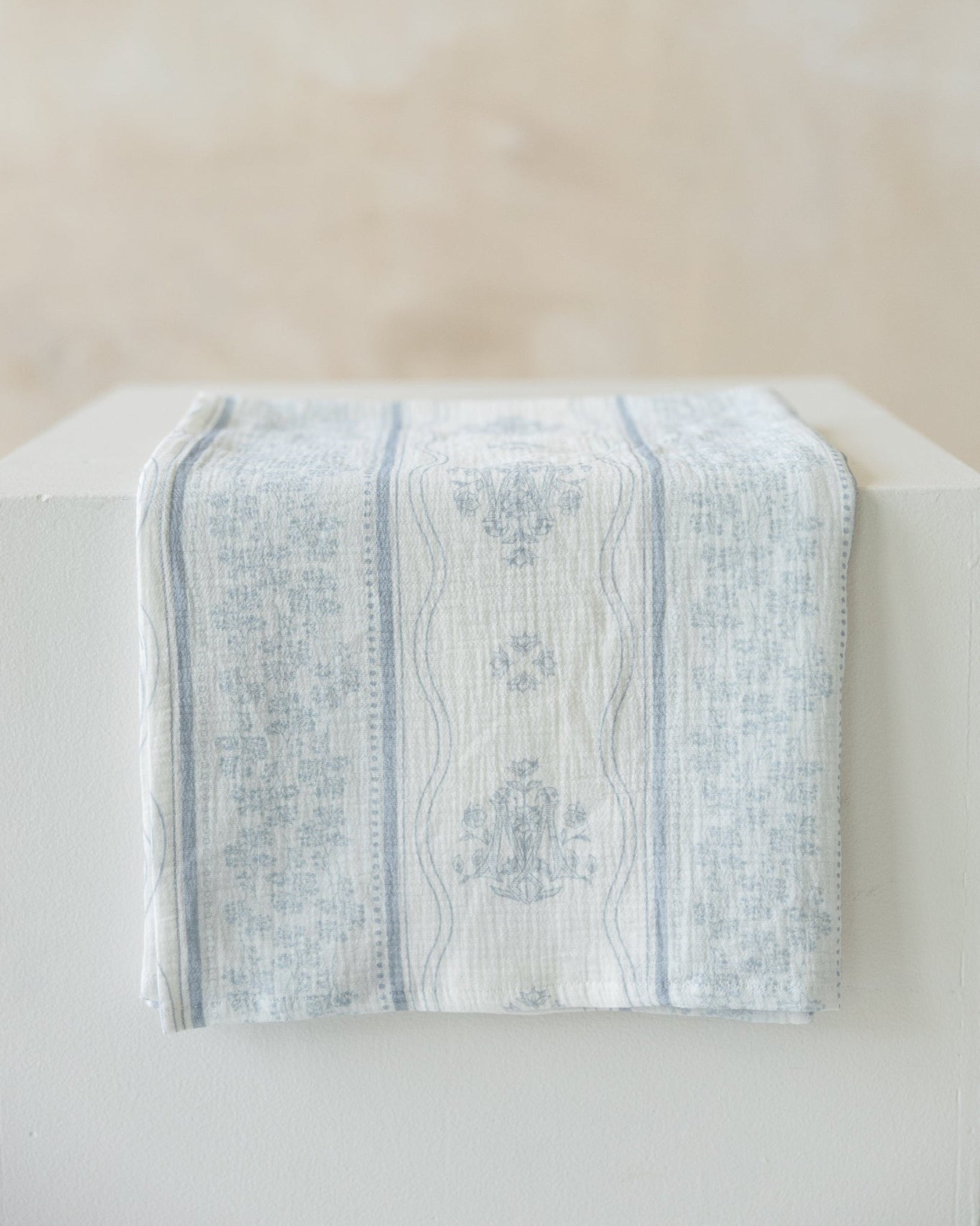 A light blue and white Marian and floral patterned cotton muslin cloth draped over a white block against a neutral beige background.