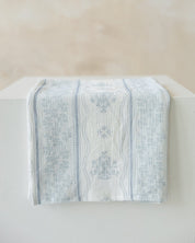 A light blue and white Marian and floral patterned cotton muslin cloth draped over a white block against a neutral beige background.