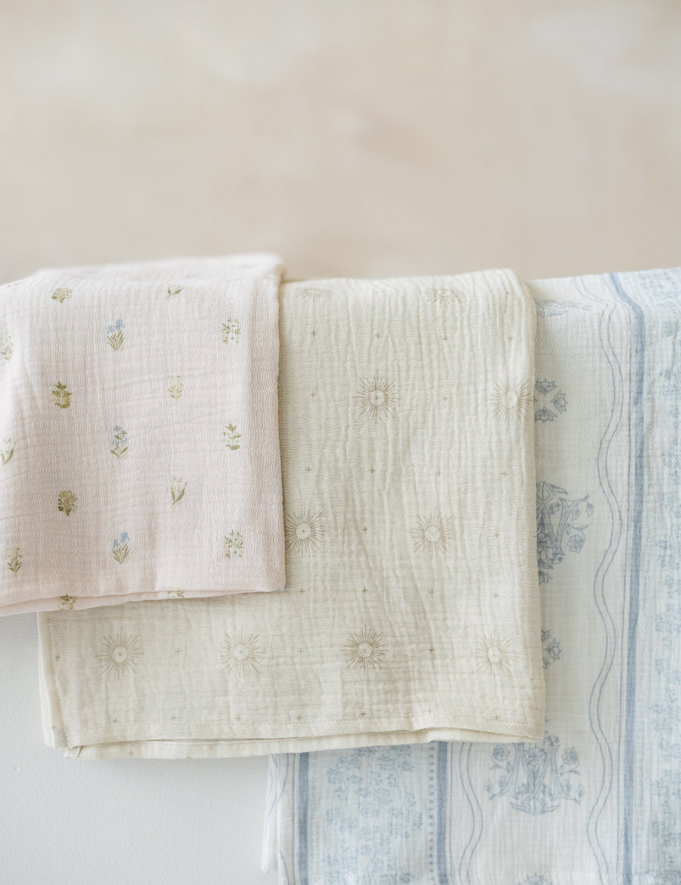 Three muslin baby swaddle blankets with catholic symbols. a blush pink one with tiny botanical florals, a sage-cream one with delicate eucharistic rays, and a white one with soft blue stripes, floral detailing, and a Marian symbol.