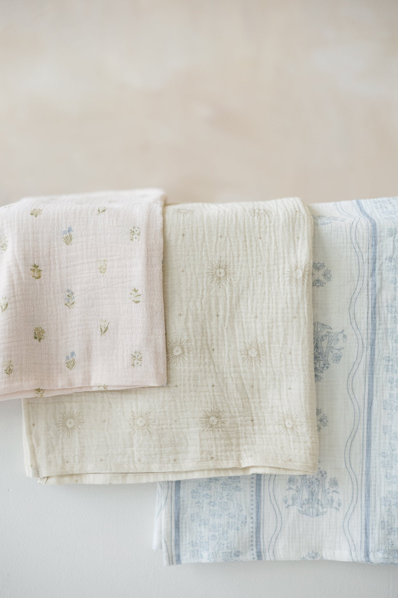 Three muslin baby swaddle blankets with catholic symbols. a blush pink one with tiny botanical florals, a sage-cream one with delicate eucharistic rays, and a white one with soft blue stripes, floral detailing, and a Marian symbol.