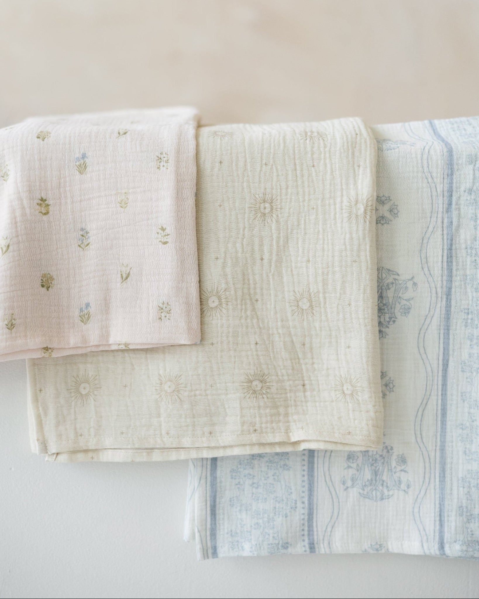 Three muslin baby swaddle blankets with catholic symbols. a blush pink one with tiny botanical florals, a sage-cream one with delicate eucharistic rays, and a white one with soft blue stripes, floral detailing, and a Marian symbol.