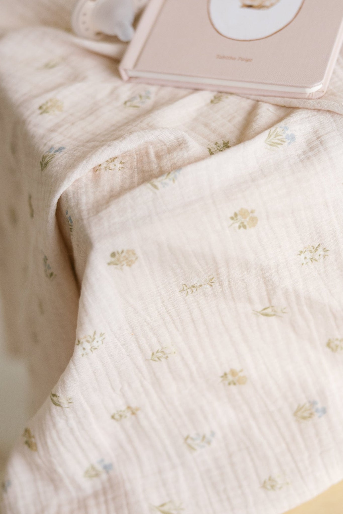 A close-up of a soft, pale blush muslin baby girl swaddle with a delicate repeating floral pattern.