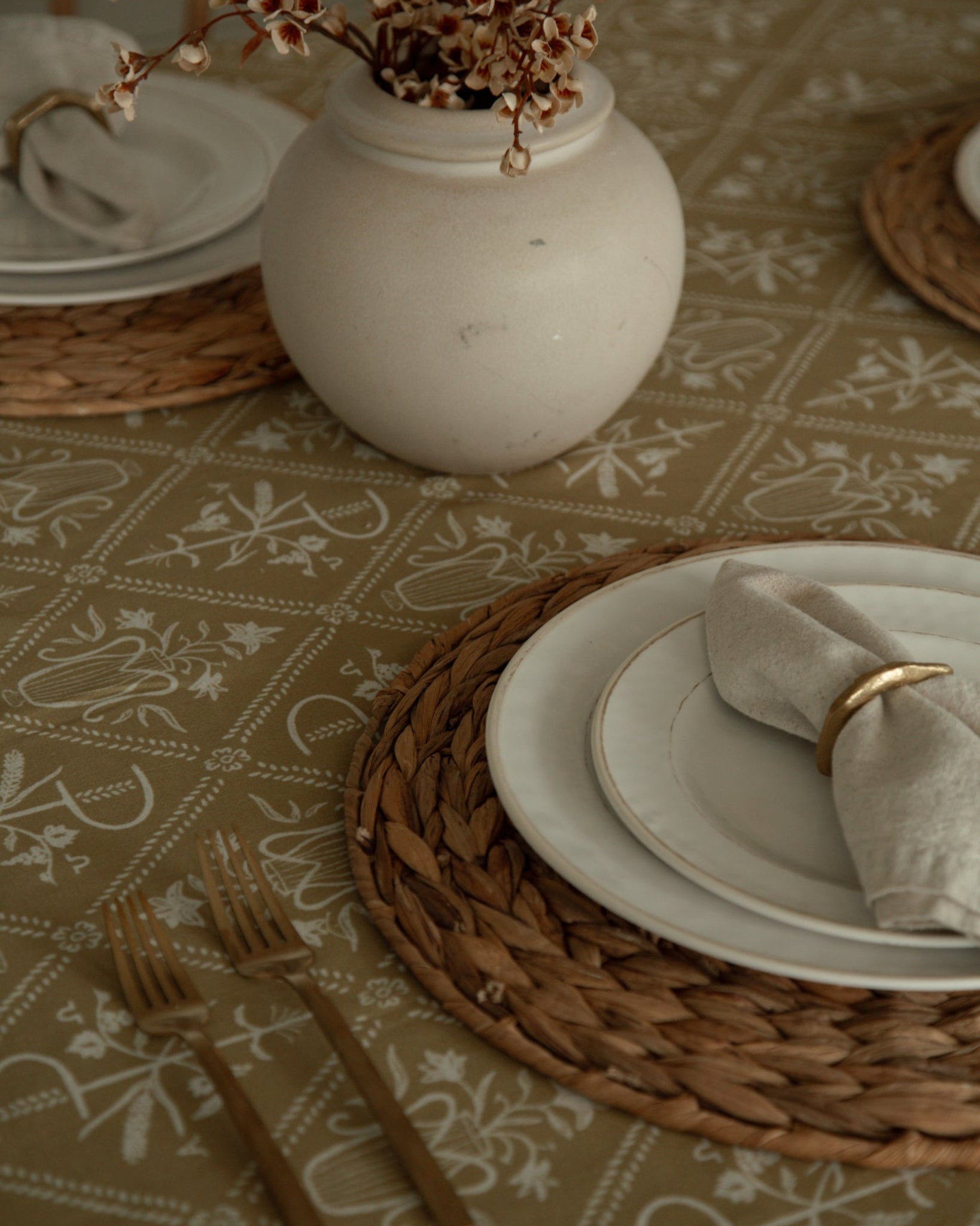A styled dining table setting featuring a beige Catholic patterned tablecloth, woven round placemats, stacked white ceramic plates, a folded cream linen napkin with a gold ring, and a white ceramic vase holding dried branches.
