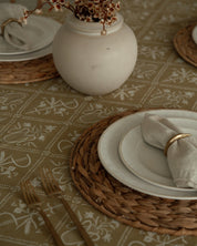A styled dining table setting featuring a beige Catholic patterned tablecloth, woven round placemats, stacked white ceramic plates, a folded cream linen napkin with a gold ring, and a white ceramic vase holding dried branches.