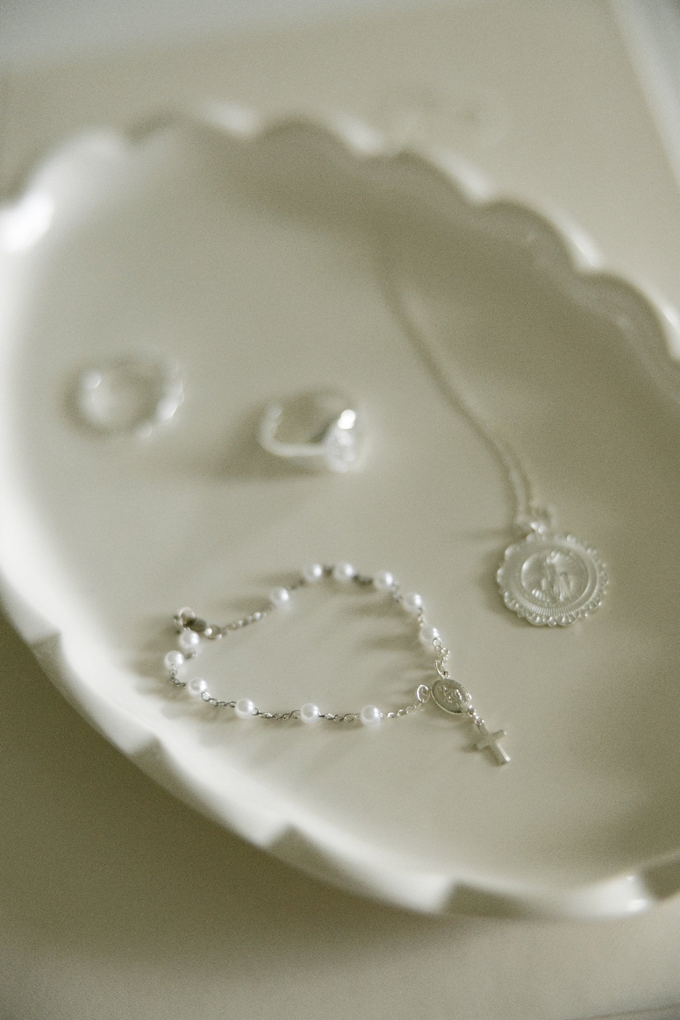 A delicate silver-toned jewelry set including a necklace with a round medallion pendant, a beaded bracelet with a small cross charm, and two rings, arranged on a scalloped white ceramic dish.