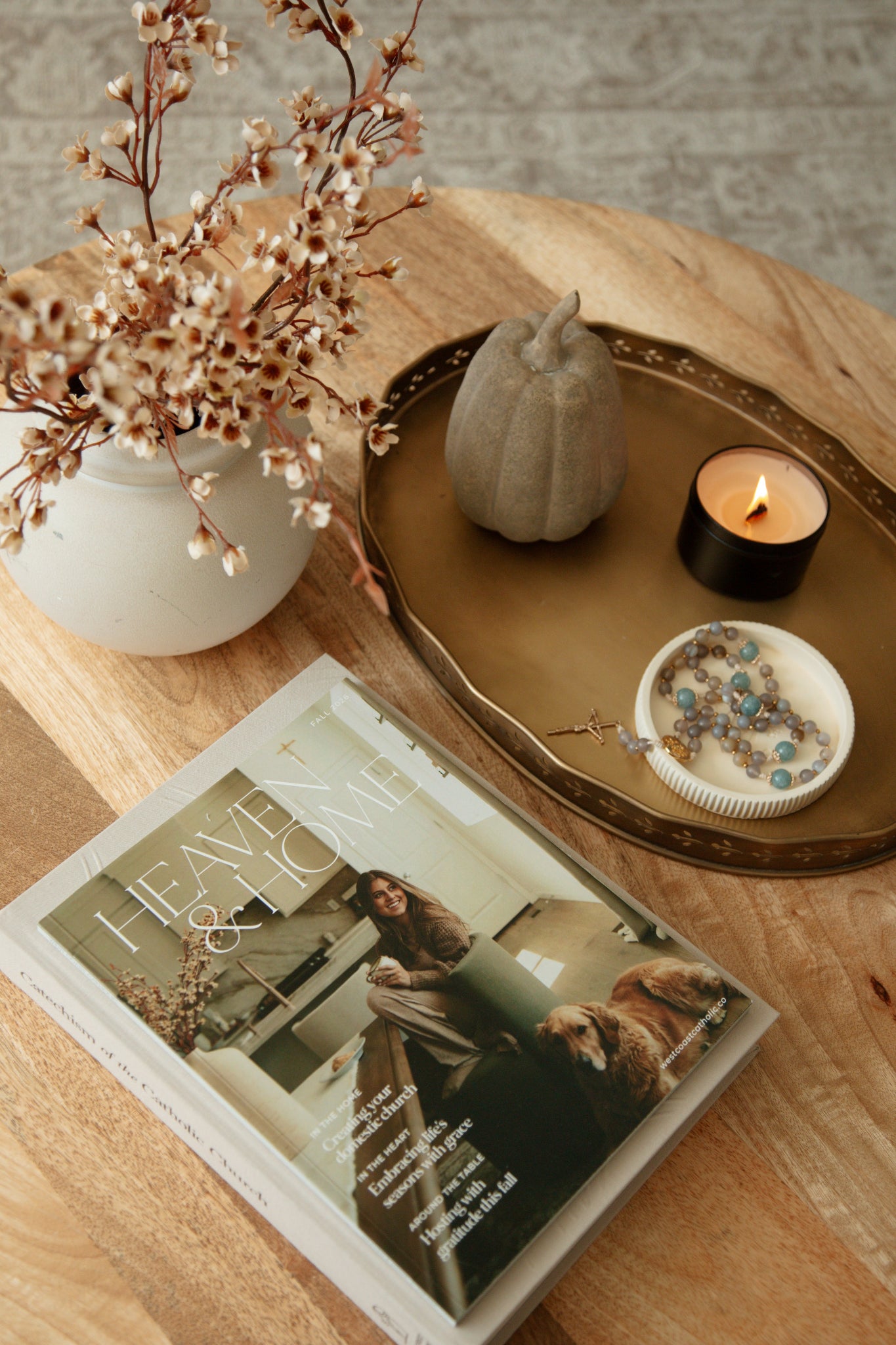 West Coast Catholic's Fall 2026 Heaven & Home magazine styled on a wooden coffee table with a lit candle, ceramic pumpkin, and a rosary for cozy Catholic fall decor.