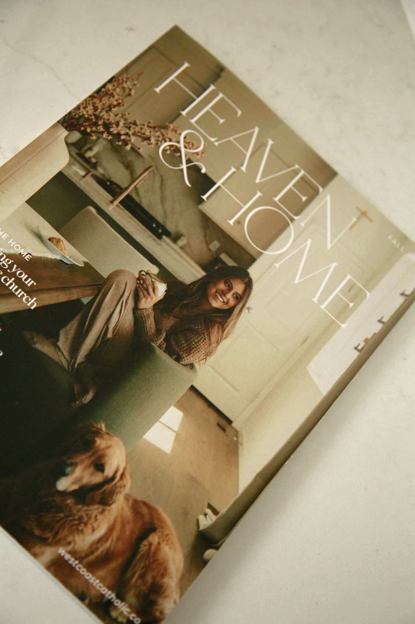 West Coast Catholic's Fall 2026 Heaven & Home magazine cover laid flat at an angle on a marble counter, featuring a woman with coffee/latte and a golden retriever.