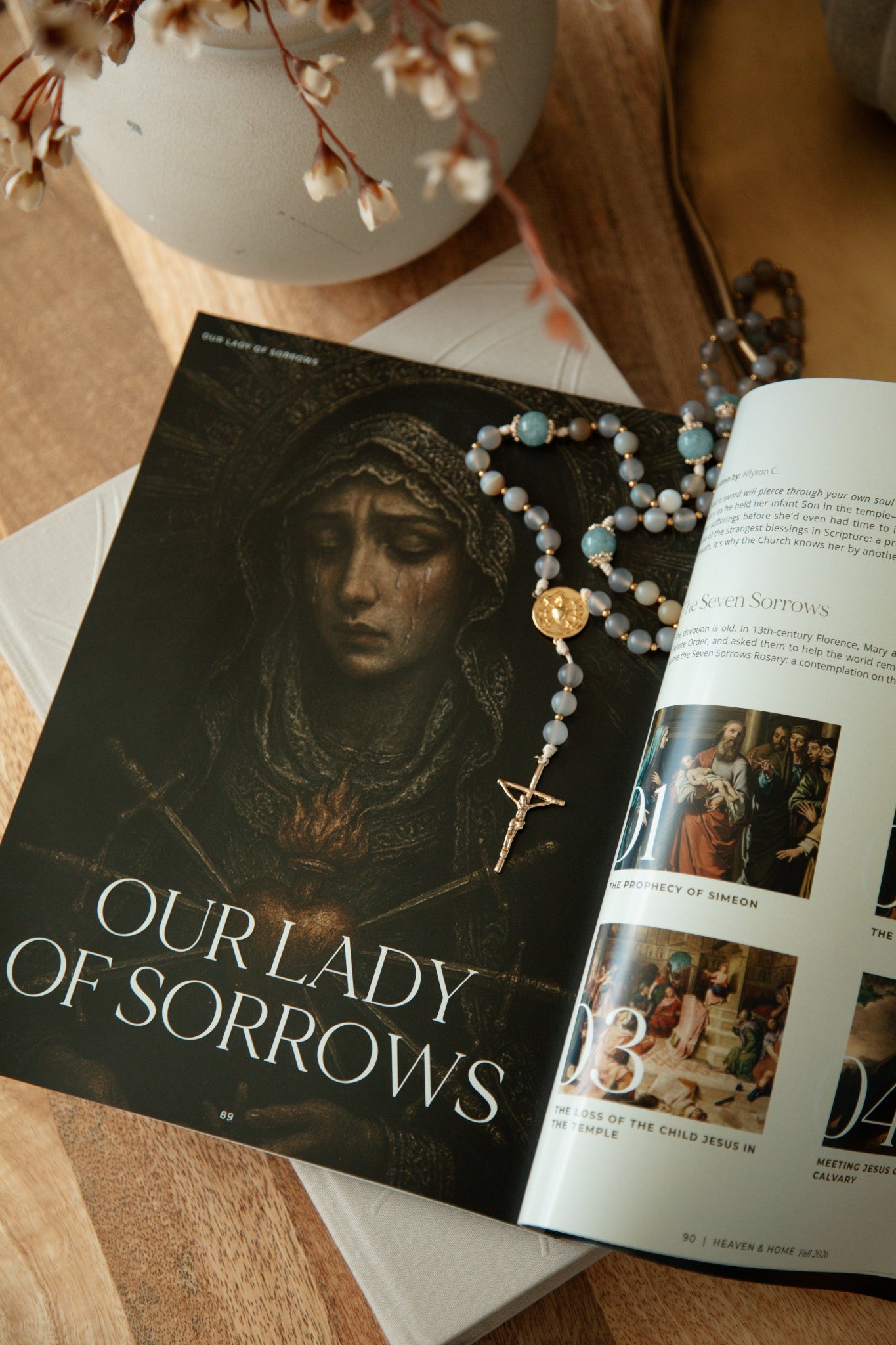 Heaven & Home magazine open to the Our Lady of Sorrows and Seven Sorrows Rosary article, styled with a blue West Coast Catholic gemstone rosary on a wooden coffee table.