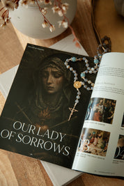 Heaven & Home magazine open to the Our Lady of Sorrows and Seven Sorrows Rosary article, styled with a blue West Coast Catholic gemstone rosary on a wooden coffee table.