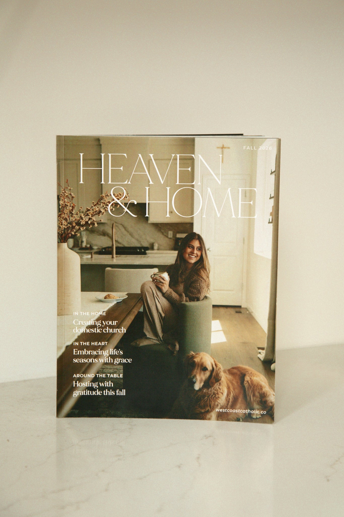 West Coast Catholic's Fall 2026 Heaven & Home magazine cover standing upright on a marble surface, featuring a woman with coffee/latte in a modern kitchen and a golden retriever.
