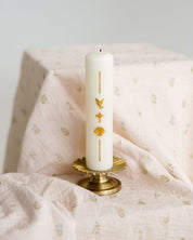 A white taper baptism candle with gold decorative symbols stands on a small gold pedestal against a soft, draped blush pink baby swaddle.