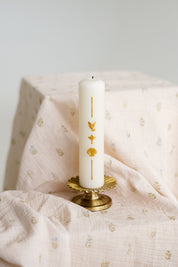 A white taper baptism candle with gold decorative symbols stands on a small gold pedestal against a soft, draped blush pink baby swaddle.
