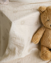 A close-up of a soft cream-colored muslin blanket with a subtle sunflower pattern, styled next to a classic brown teddy bear.