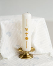 A white taper candle with gold religious symbols (dove, cross, anchor, shell) on its side, displayed in a gold metal holder on a patterned white fabric surface.