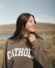 Woman wearing a brown sweatshirt with 'CATHOLI' text, standing outdoors with a scenic background.