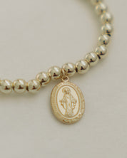 Beaded gold bracelet with a Miraculous Medal on a light gray background. 