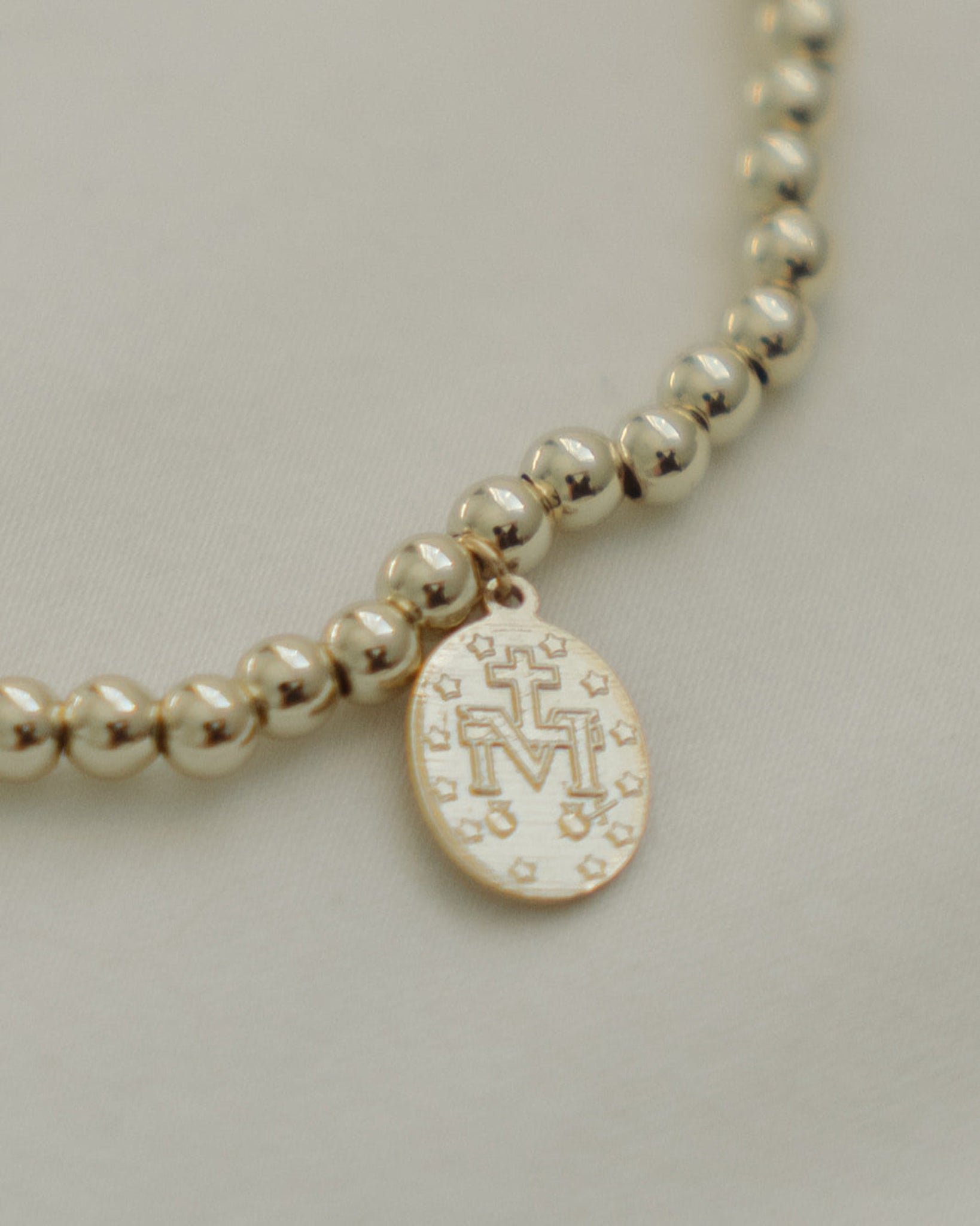 Gold beaded necklace with a round pendant with a Marian symbol on a light gray background