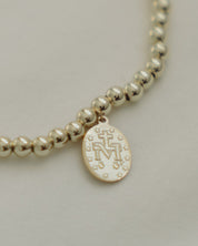 Gold beaded necklace with a round pendant with a Marian symbol on a light gray background