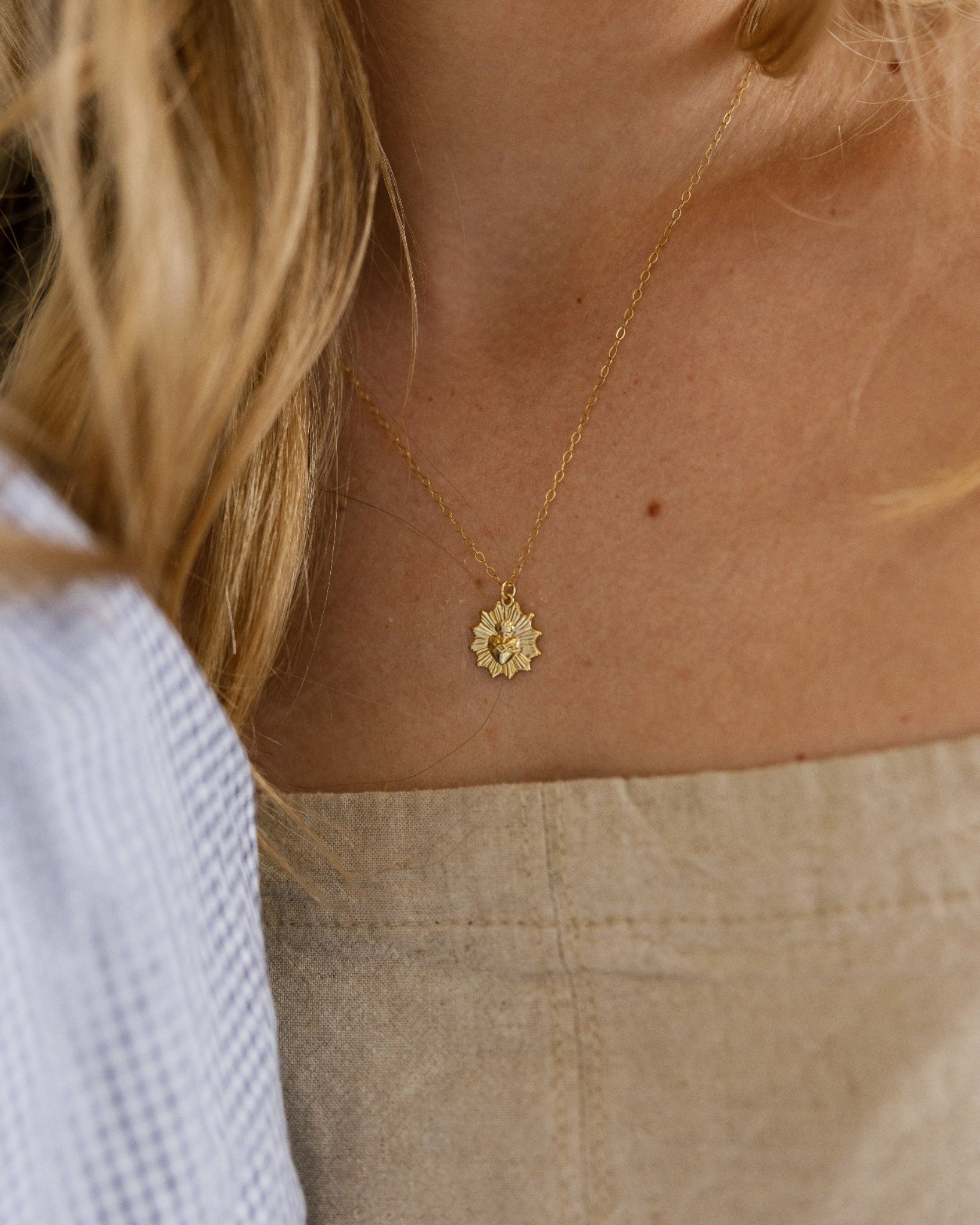 A close-up lifestyle shot of a woman wearing a delicate gold chain necklace with a small, sacred heart pendent. She is wearing a beige linen top.