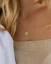 A close-up lifestyle shot of a woman wearing a delicate gold chain necklace with a small, sacred heart pendent. She is wearing a beige linen top.