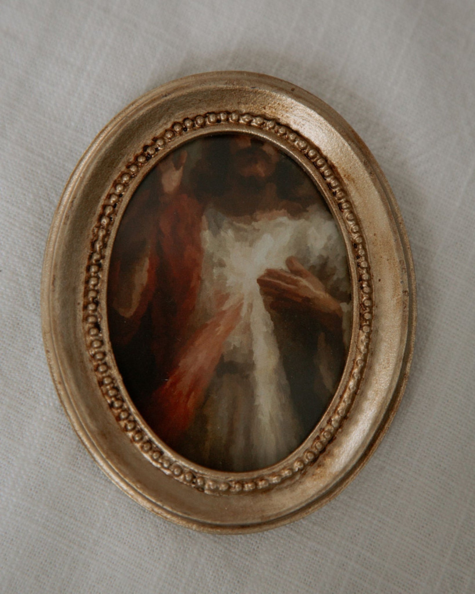 Small oval catholic painting in a decorative gold frame on a textured beige wall.