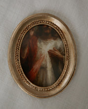 Small oval catholic painting in a decorative gold frame on a textured beige wall.