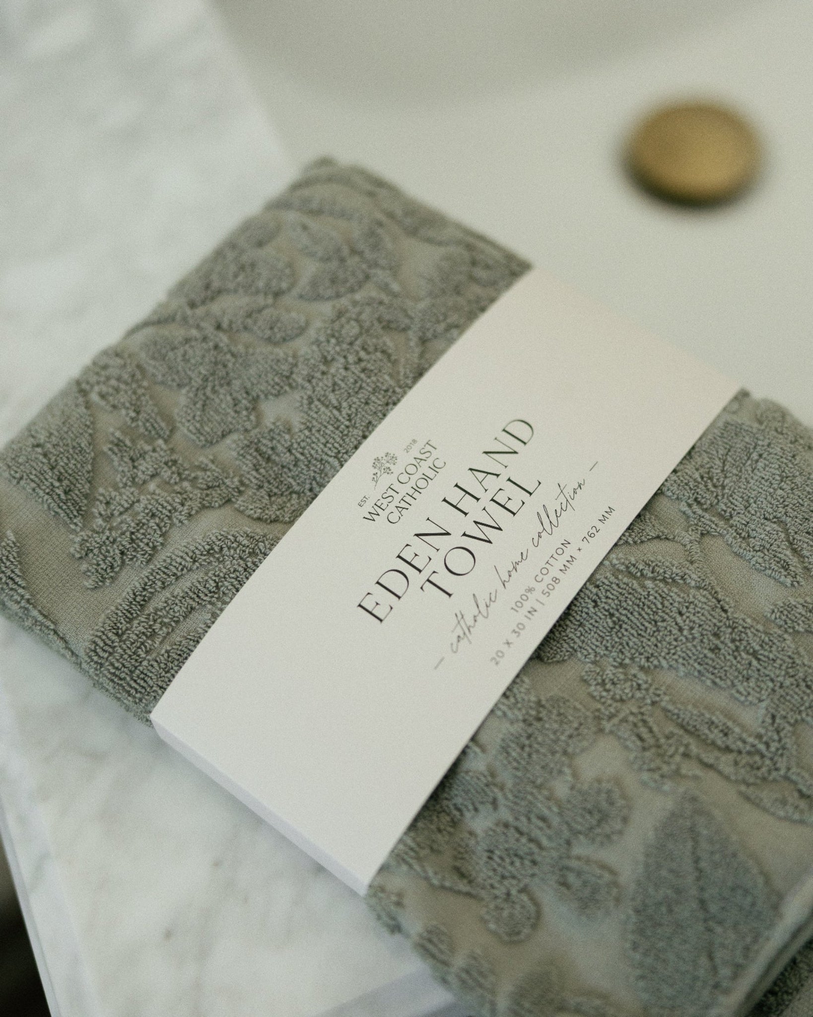 A folded gray textured hand towel with a white paper label reading 'EDEN HAND TOWEL' and 'The Handcrafted Collection,' placed on a white marble surface near a sink.