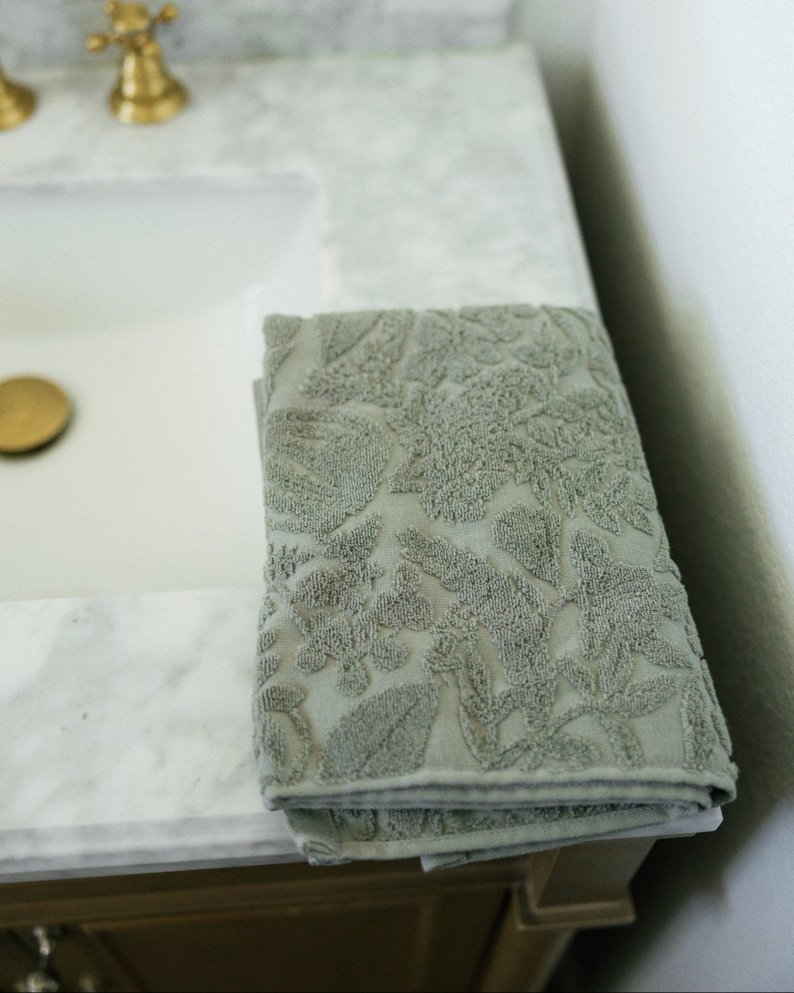 A folded gray textured bath towel with a raised floral pattern rests on a white marble bathroom vanity countertop next to a sink with gold fixtures.