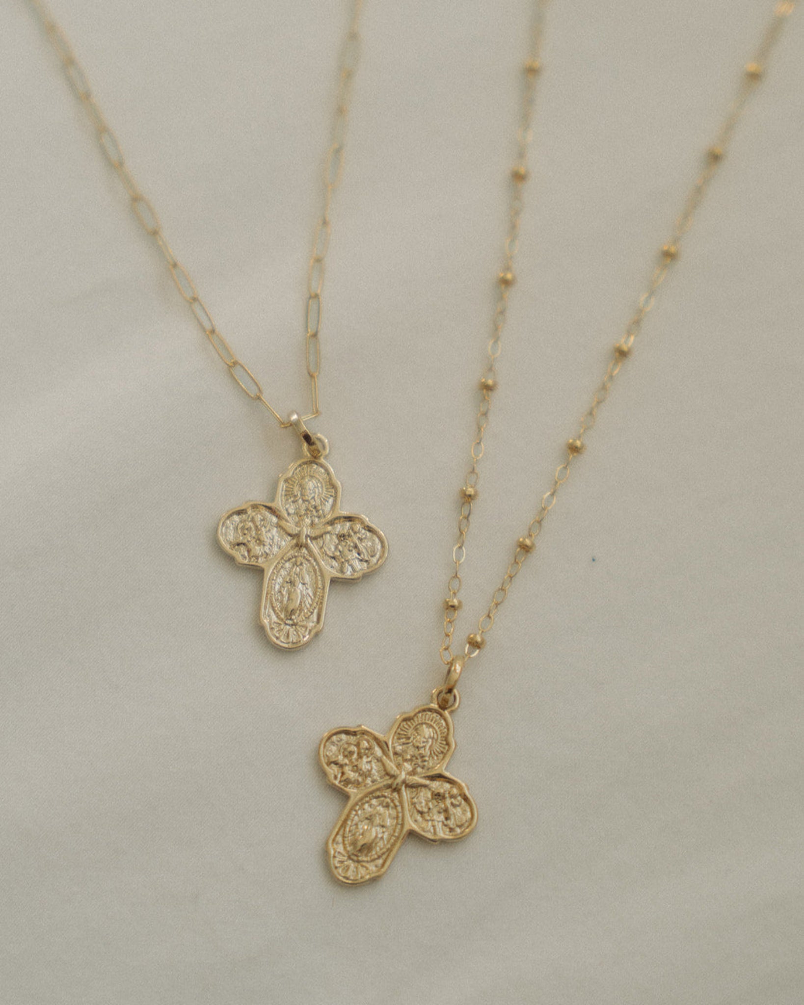 Two four-way gold cross necklaces, one with a beaded chain and one with a paperclip chain. Depicted on the crosses are the Holy Trinity and Mary. All lying on a white-cream surface.