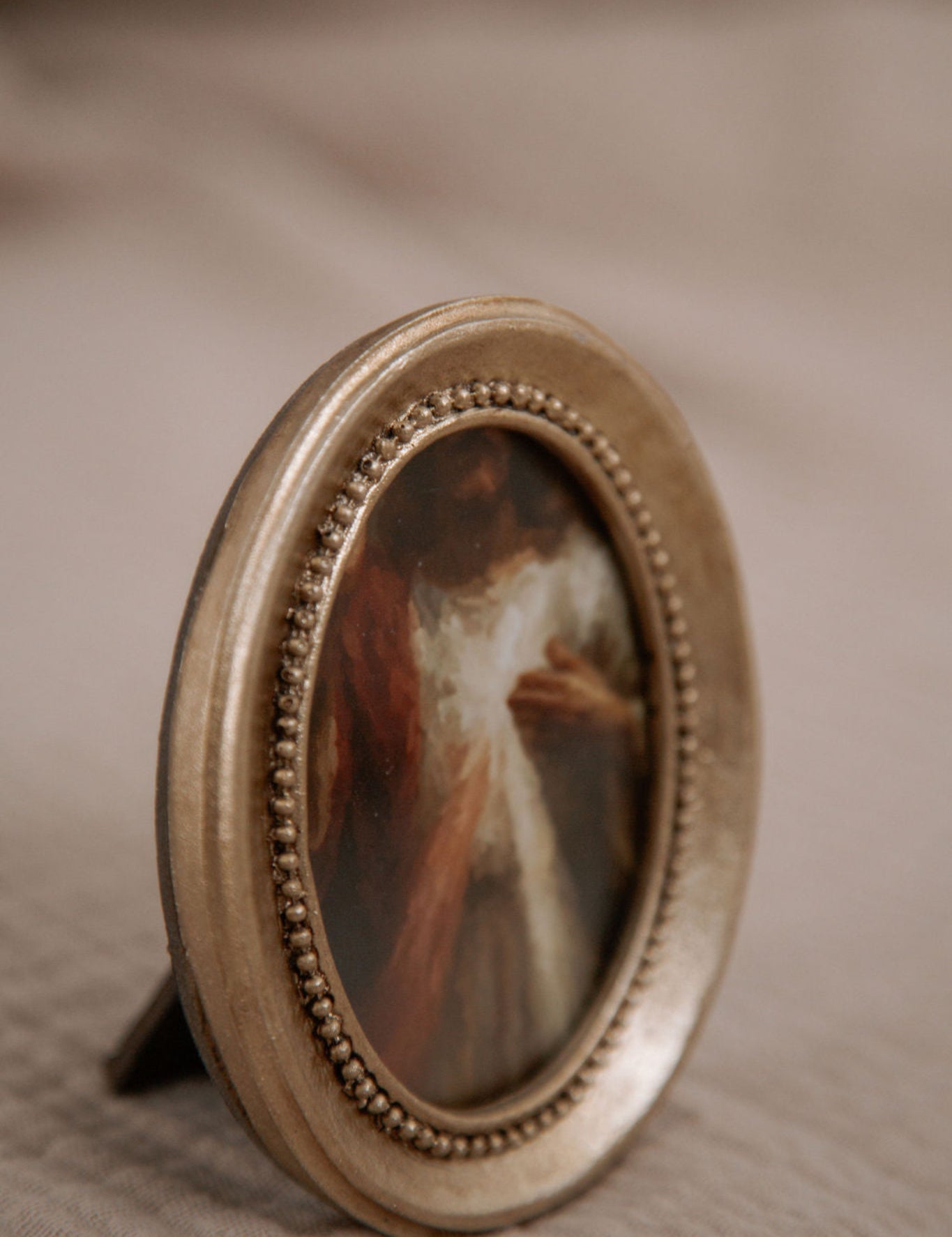 Round gold-framed miniature painting of the Divine Mercy on a textured beige surface