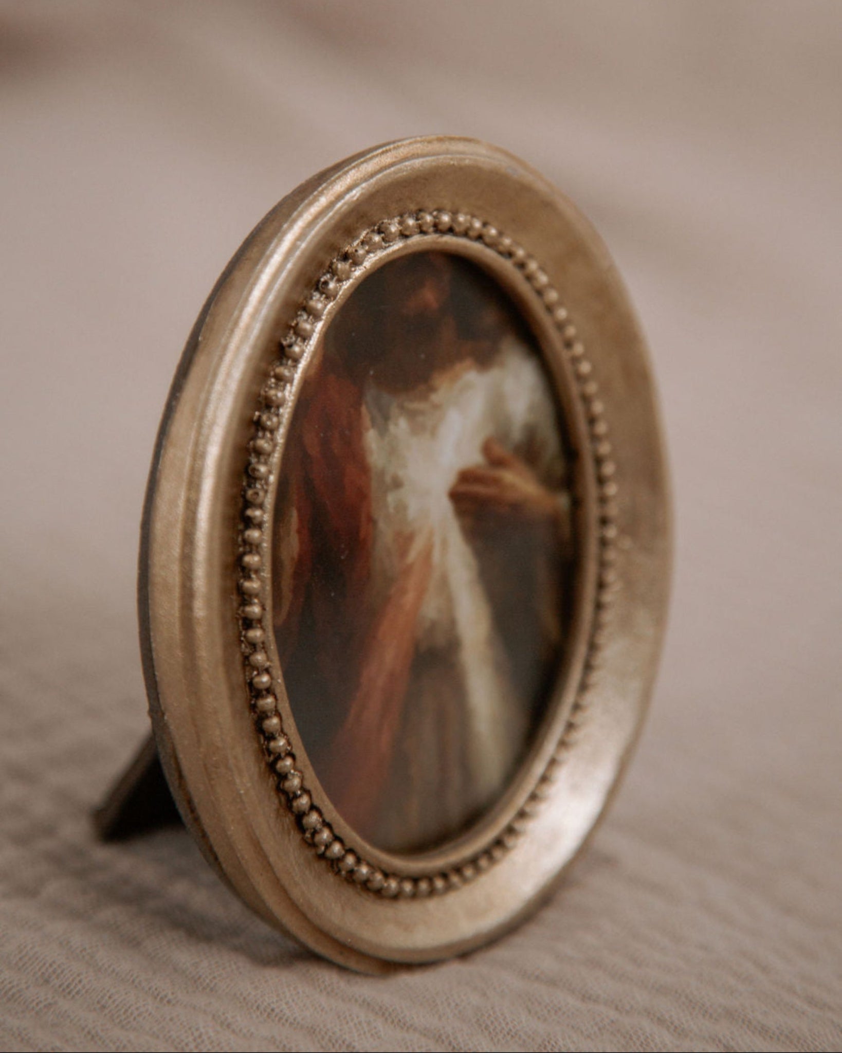 Round gold-framed miniature painting of the Divine Mercy on a textured beige surface