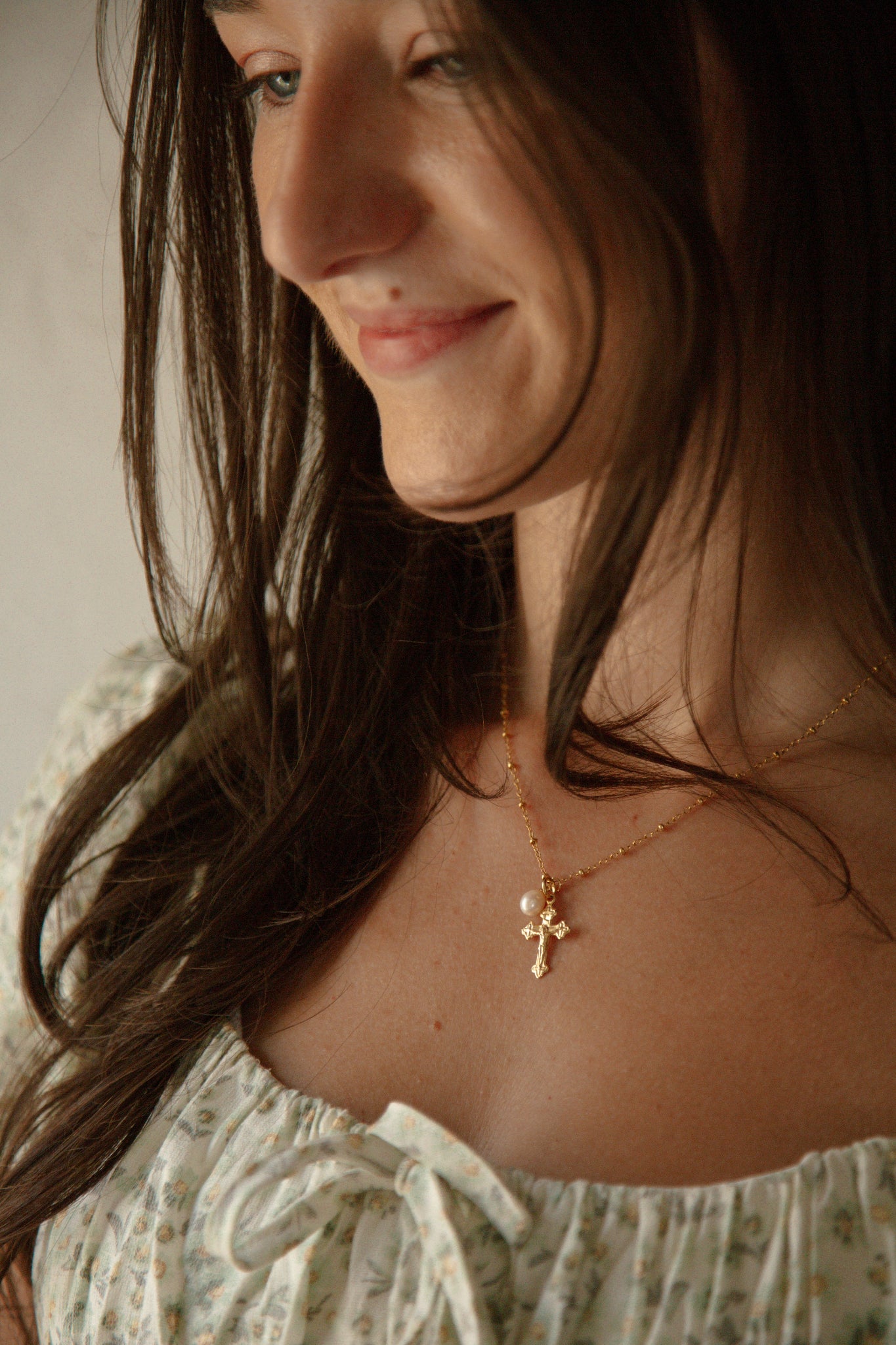 A close-up lifestyle shot of a woman wearing a delicate gold crucifix necklace with small white pearl, styled with a floral top.