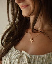 A close-up lifestyle shot of a woman wearing a delicate gold crucifix necklace with small white pearl, styled with a floral top.