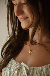 A close-up lifestyle shot of a woman wearing a delicate gold crucifix necklace with small white pearl, styled with a floral top.