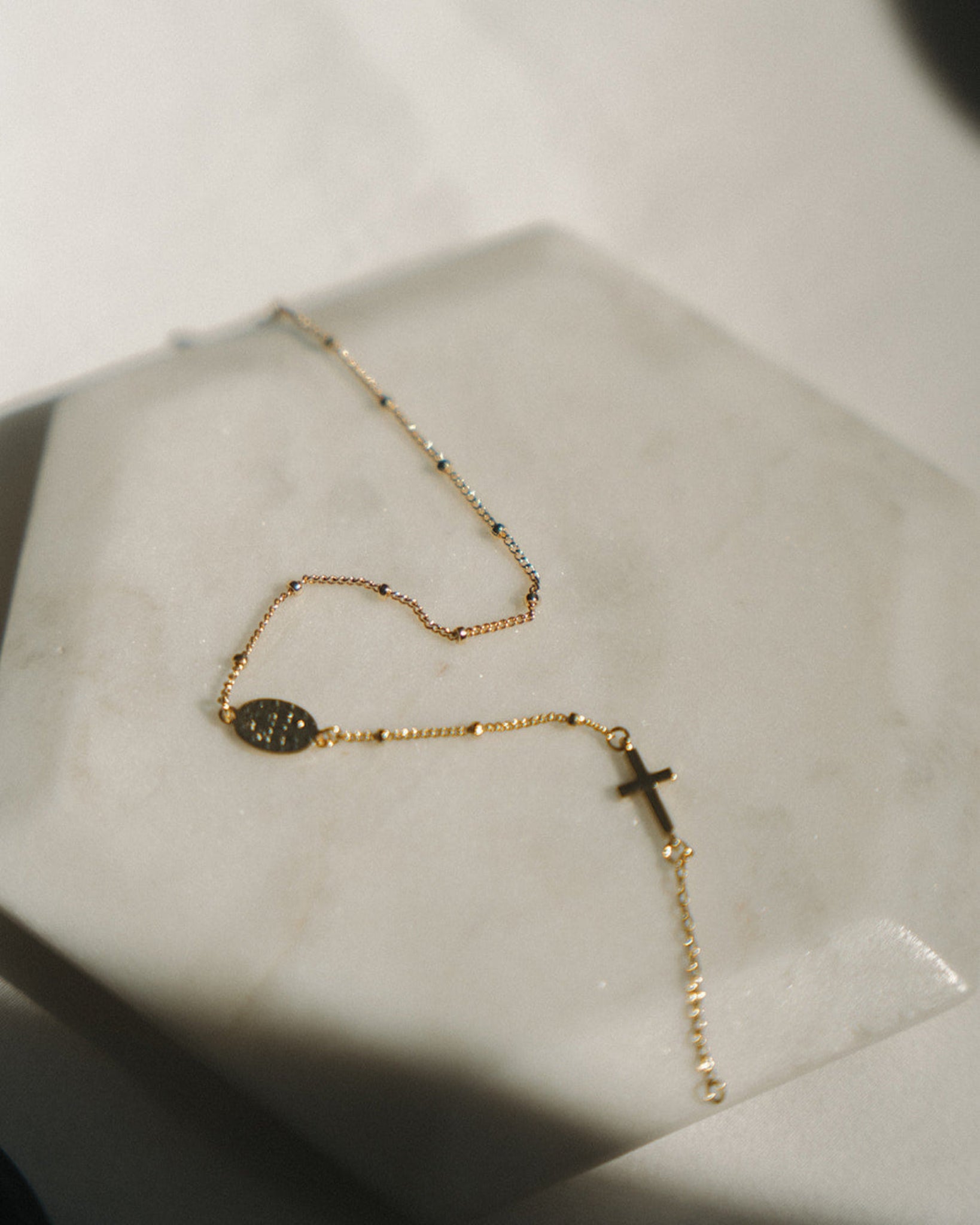 Gold dainty rosary bracelet with a cross pendant on a light gray surface